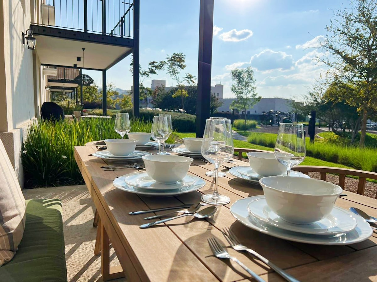 An outdoor dining table is set for a meal, featuring white dishware and glassware arranged neatly. The table is surrounded by greenery and sunlight, creating a bright and inviting ambiance. Comfortable seating is visible in the background, enhancing the outdoor experience.