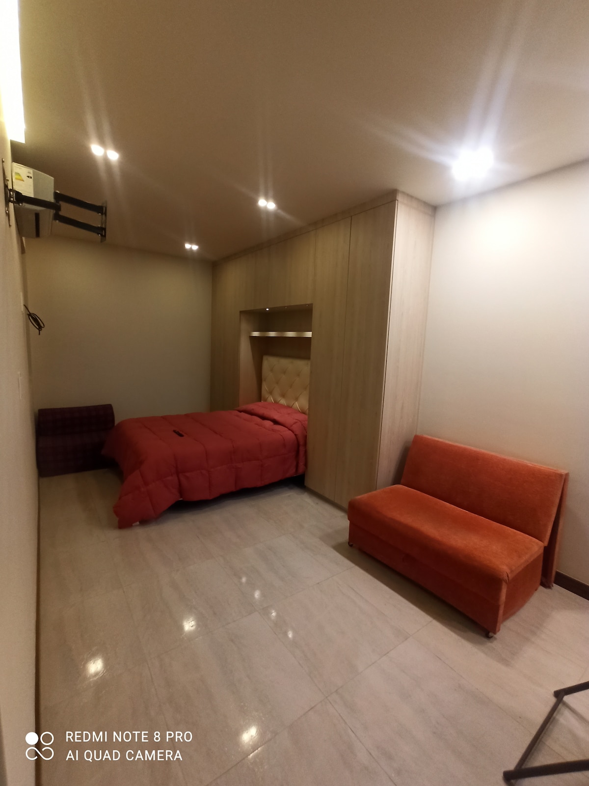 A cozy room is presented with warm lighting, featuring a spacious bed with a red coverlet. A plush orange sofa is positioned in the corner, enhancing the inviting ambiance. Smooth, light-colored flooring complements the wooden paneling on the wall, contributing to a contemporary design.