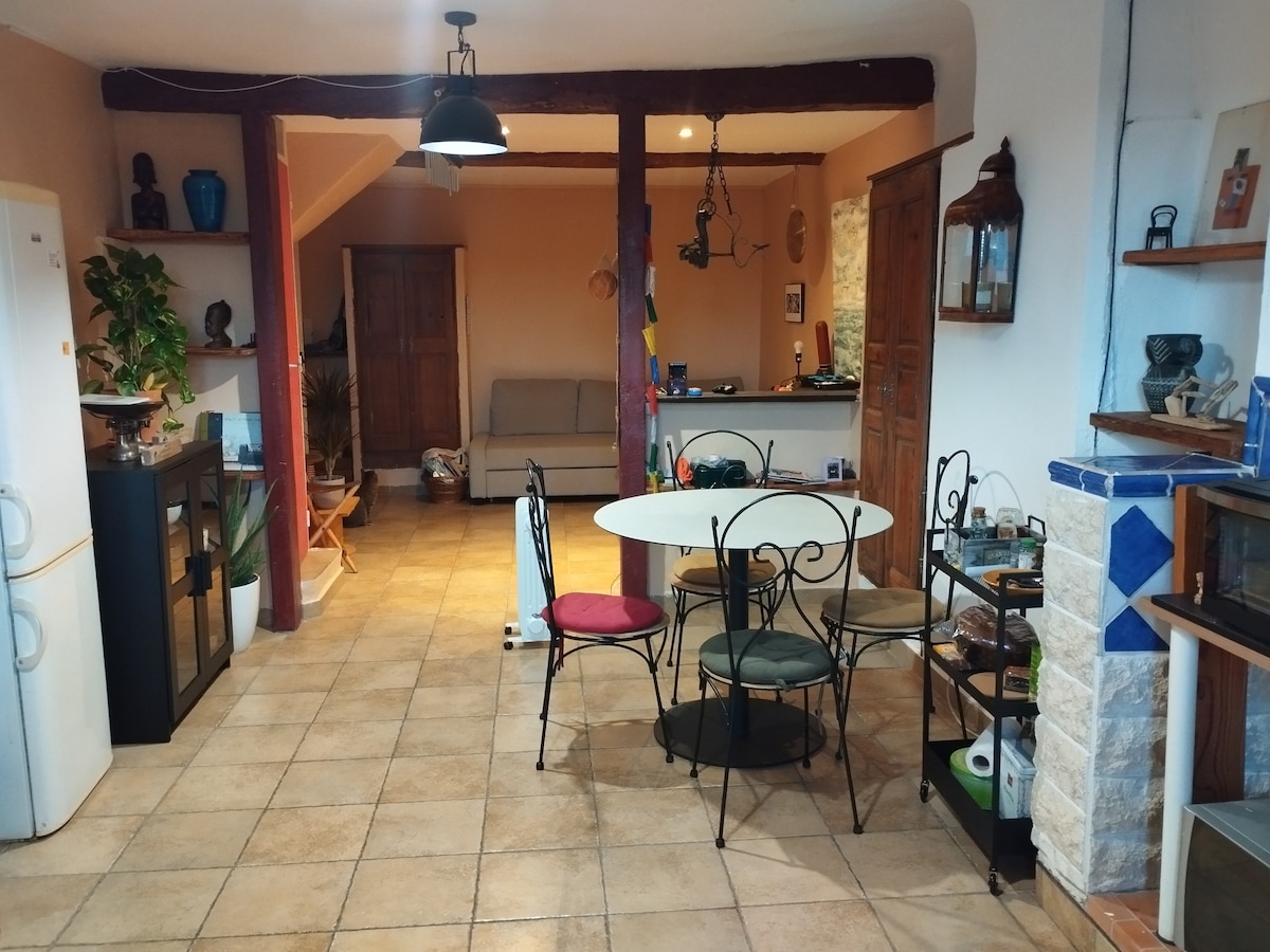 A spacious dining area is presented with a round table and four chairs, two of which are adorned in vibrant colors. An open kitchen is visible, featuring appliances like a refrigerator. Natural light fills the room, with a cozy living space seen in the background.