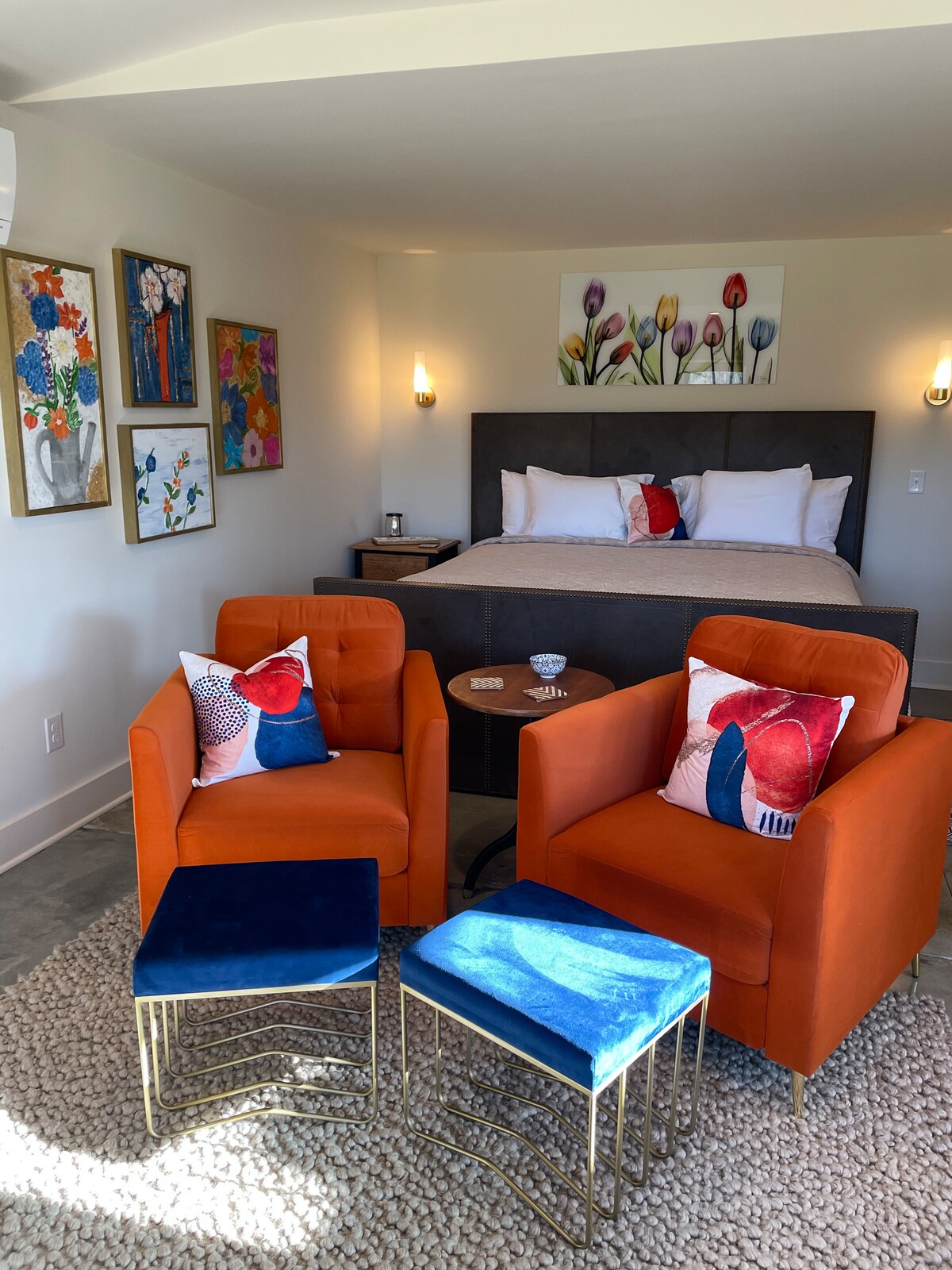 A cozy studio space features a king-sized bed against the wall, adorned with floral artwork. Two orange chairs with patterned cushions sit across from a small table, complemented by colorful ottomans. Soft lighting enhances the inviting atmosphere while area rugs add texture to the polished concrete floor.