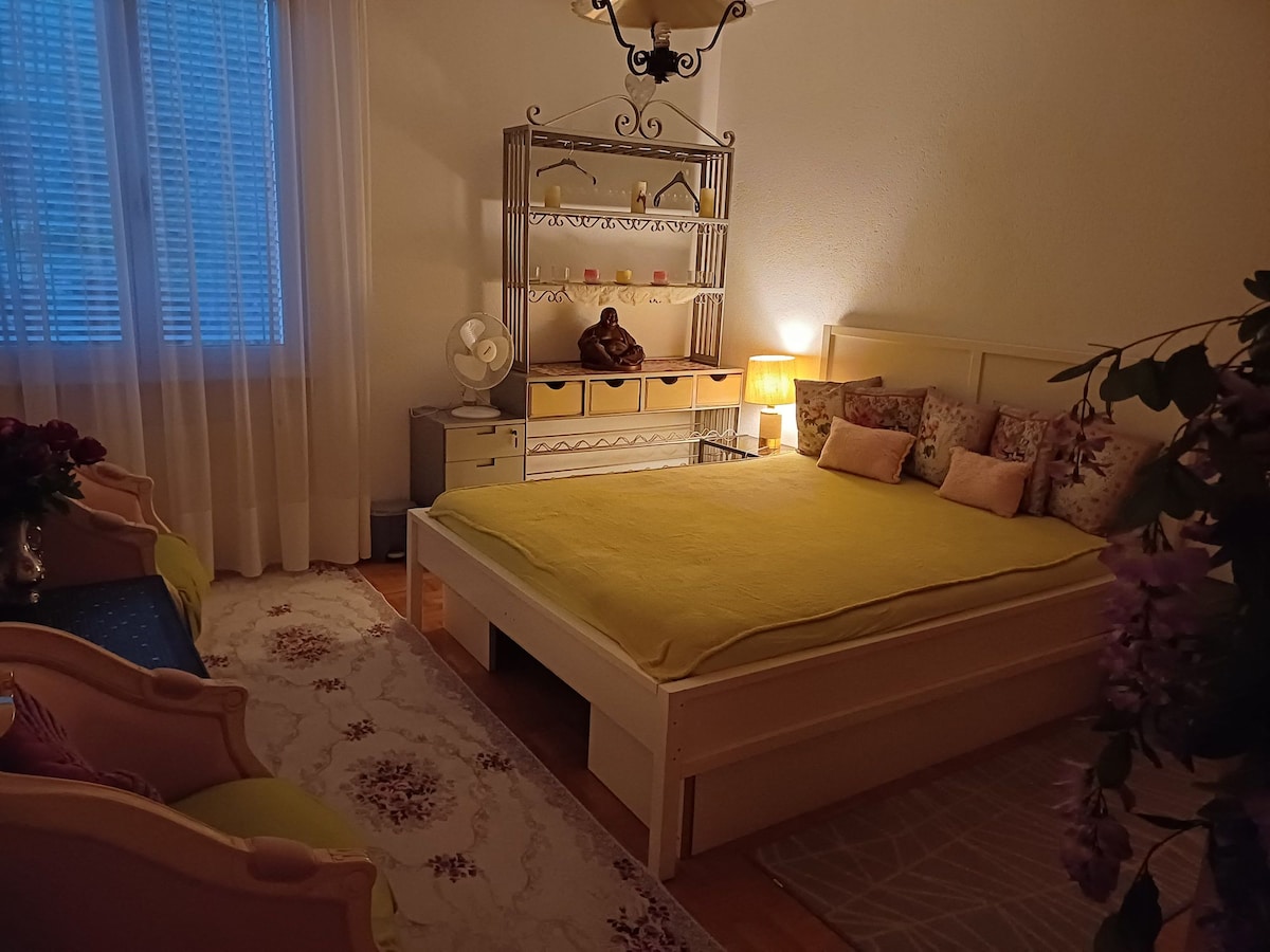 A cozy sleeping area features a spacious bed with a yellow blanket casually draped over it. Decorative pillows add a touch of comfort. Soft lighting from a bedside lamp creates a serene atmosphere, while a fan is positioned nearby. A decorative shelf and two armchairs complete the inviting space.