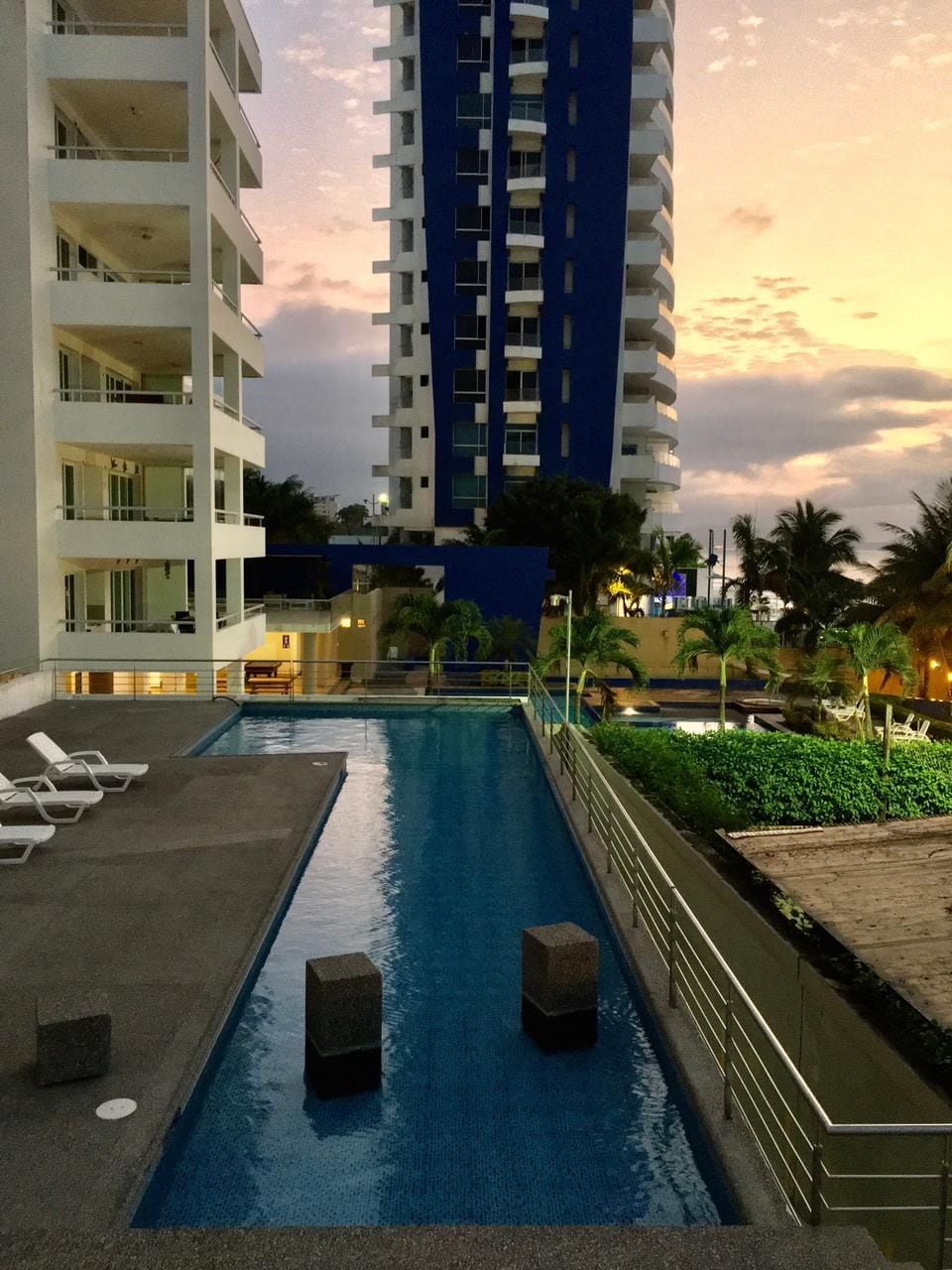 A serene pool area is featured, reflecting the twilight sky. Comfortable lounge chairs are placed along the pool's edge. Lush greenery and palm trees surround the landscape, with a modern building visible in the background, enhancing the relaxed atmosphere of the space.