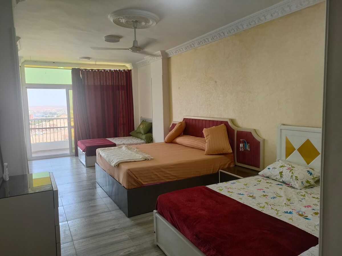 A spacious room is furnished with a large bed and a smaller bed, both positioned for comfort. Natural light enters through a large window, creating a bright environment. Decorative cushions add color to the bedding. The room offers access to a balcony with views.