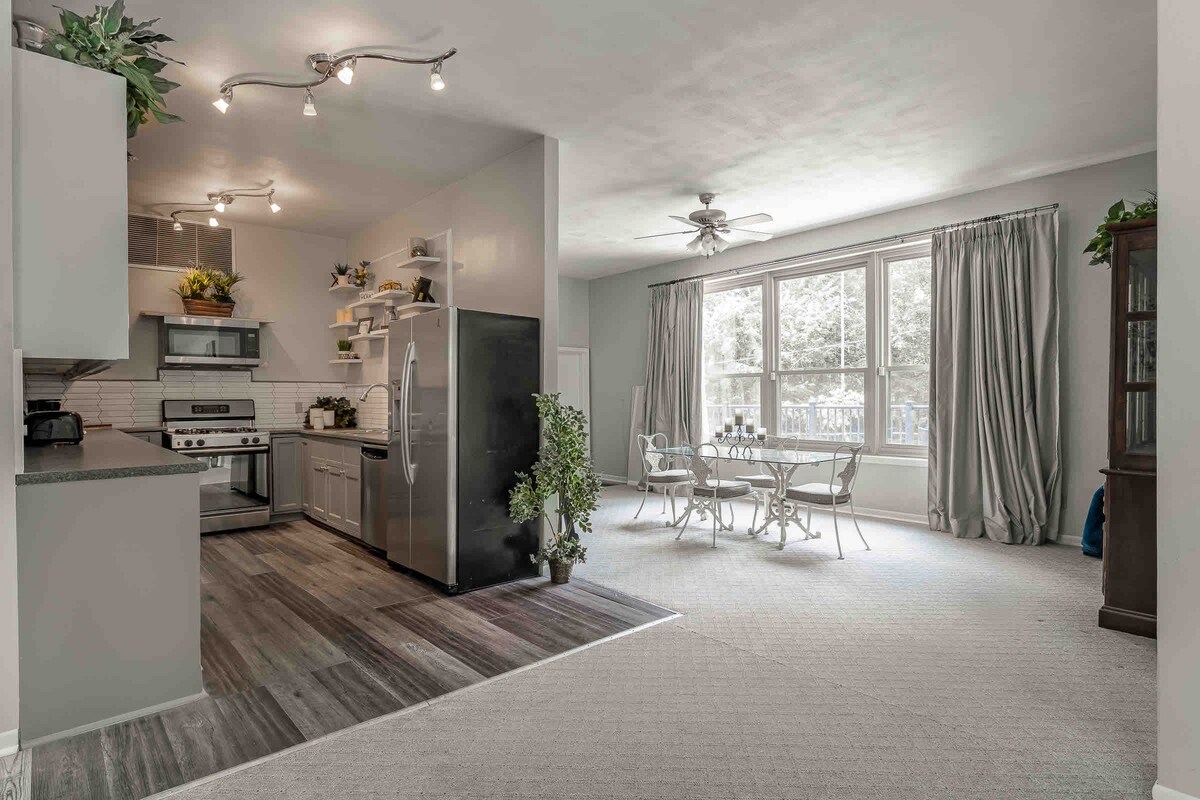 An open layout kitchen and dining area is displayed, featuring modern appliances and ample counter space. Two large windows provide natural light, while a dining table with four chairs sits adjacent to the kitchen. Soft carpet enhances the spacious feeling of the room.