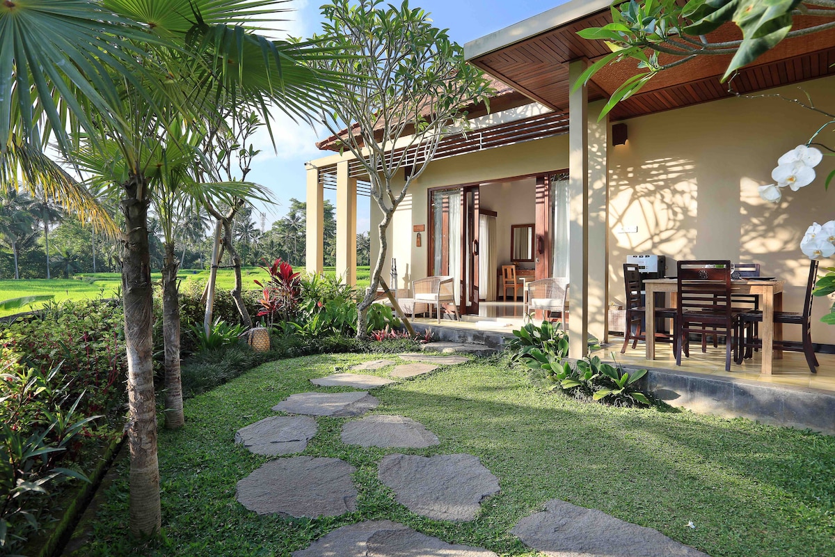 A pathway of natural stone leads to a spacious porch area, framed by lush greenery and tropical plants. Outdoor dining furniture is visible under the covered area, providing views of the surrounding rice fields. The peaceful environment highlights the villa's connection to nature.