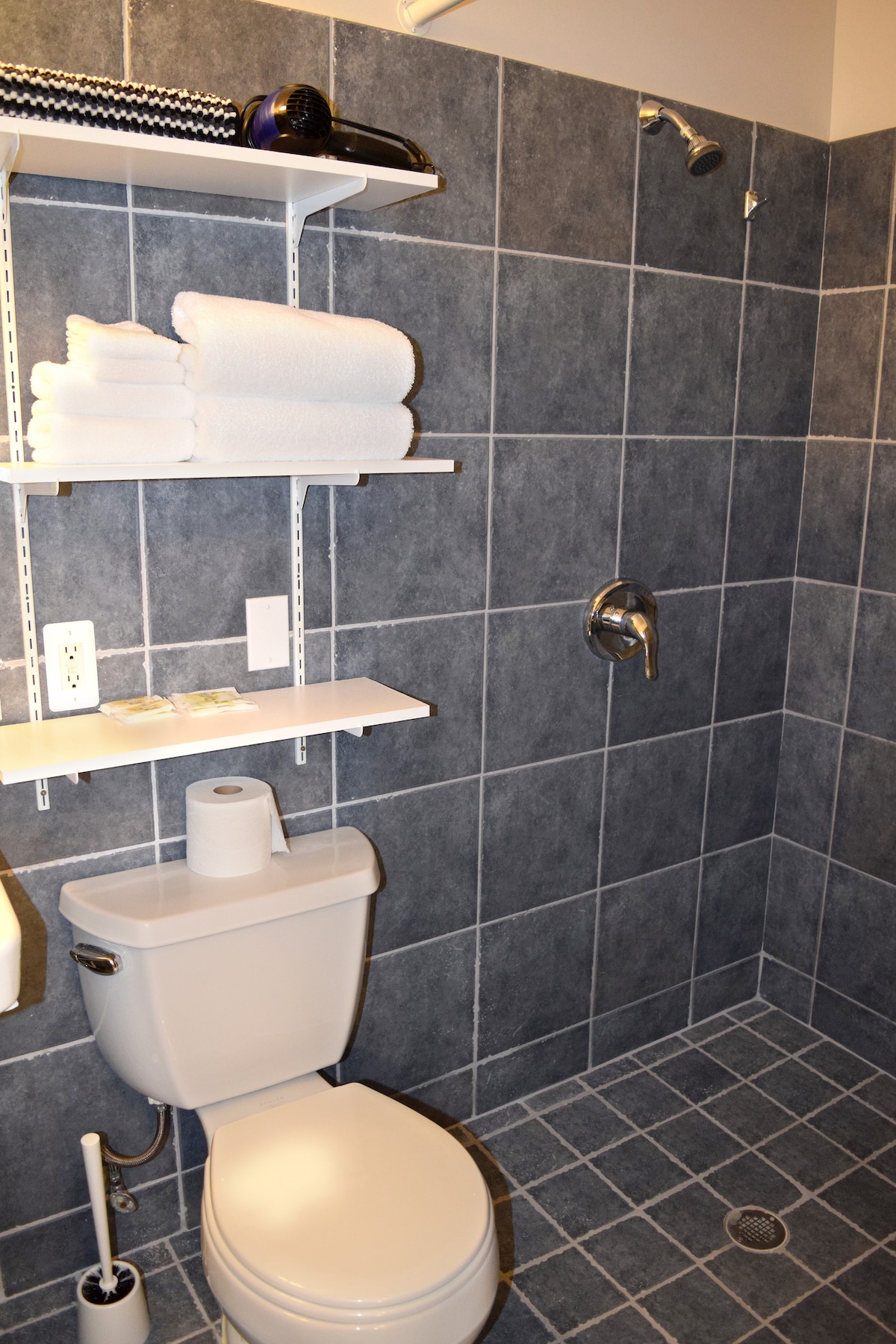 A modern bathroom is shown, featuring gray tiled walls and floor. Shelves hold neatly stacked white towels, with toiletries placed on the counter. A white toilet sits adjacent to the shower, which has an overhead water fixture.