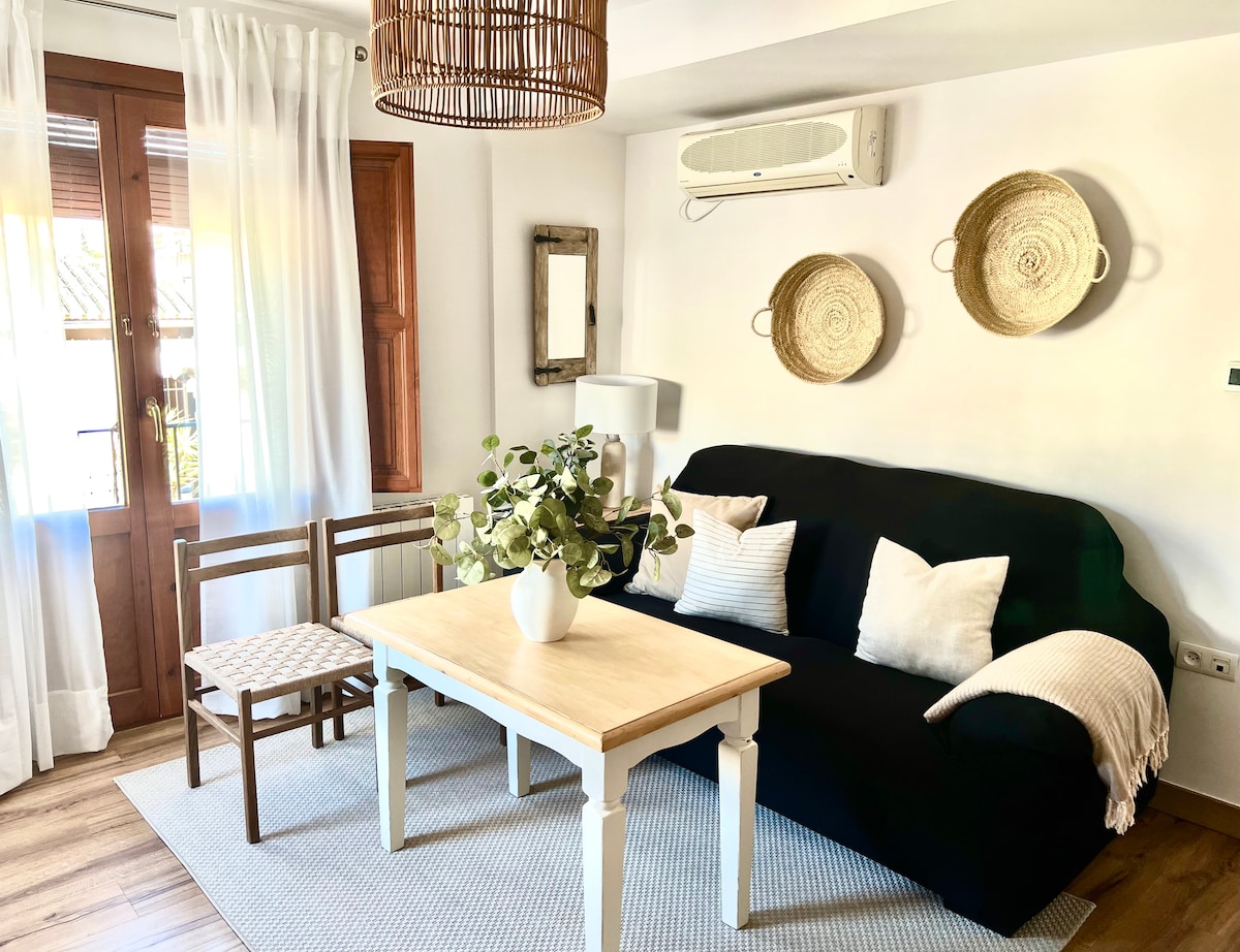 A cozy living area is furnished with a dark sofa and a wooden table. Two chairs sit beside the table, and a vase with greenery adds a touch of nature. The walls are decorated with woven wall art, and natural light filters through the white curtains.
