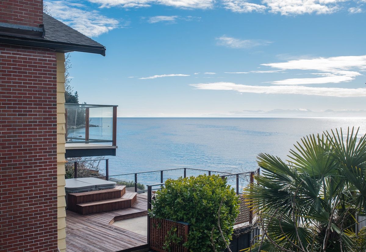 A private deck is seen featuring a hot tub surrounded by lush greenery, overlooking the tranquil ocean. The vast sea stretches towards the horizon under a clear blue sky, while the brick exterior of the house provides a sturdy, warm contrast to the scenery.