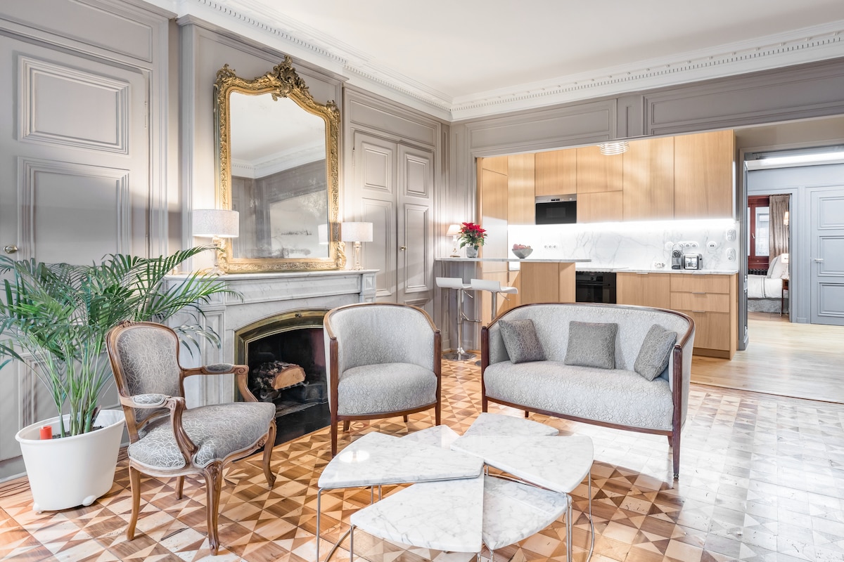 The living area features elegant gray walls adorned with a decorative mirror, complemented by two upholstered chairs and a marble-topped coffee table. A cozy fireplace is situated against one wall, while an open kitchen with wooden cabinetry is visible in the background.
