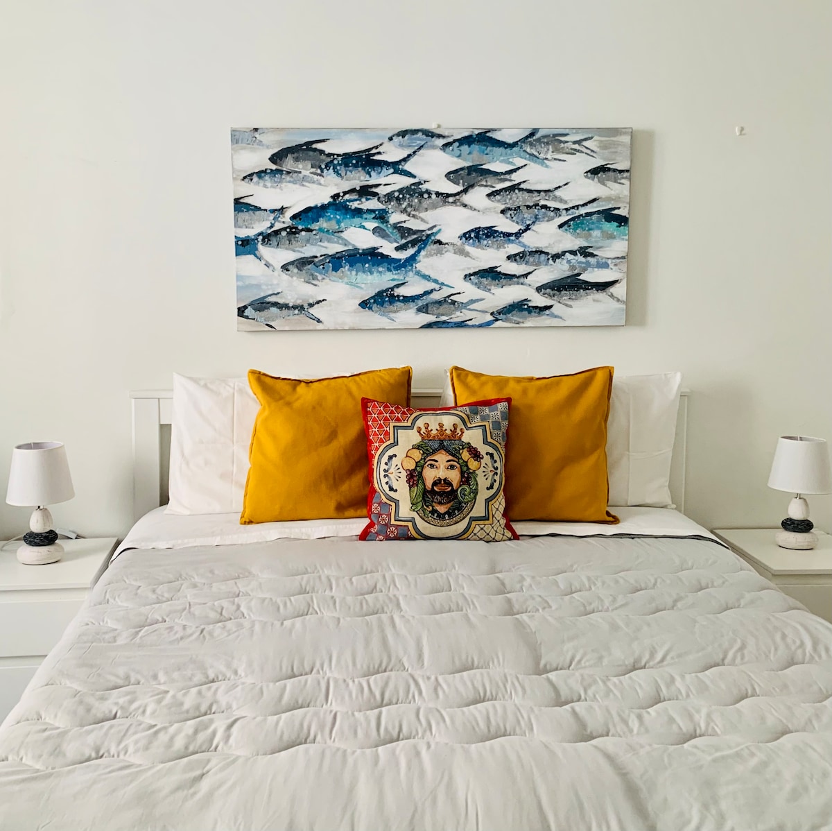 A sleeping area features a spacious bed adorned with a light quilt and bright yellow accent pillows. A decorative throw pillow adds a pop of color. Two bedside lamps are situated on either side of the bed, and a coastal-themed artwork hangs above the headboard.