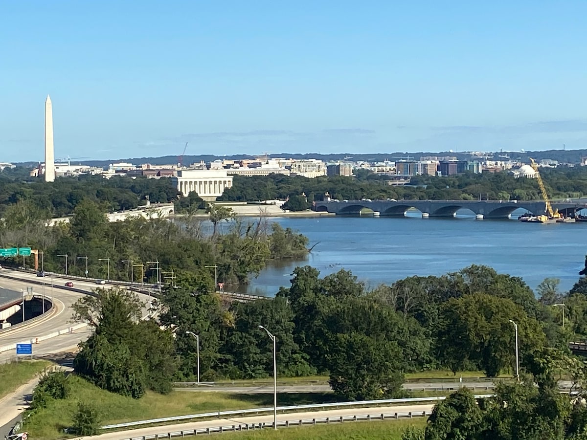 A clear view showcases the Washington Monument and the Lincoln Memorial across the Potomac River. Lush greenery lines the riverbanks, and bridges connect the areas in the foreground. The vibrant blue sky enhances the scenic landscape.