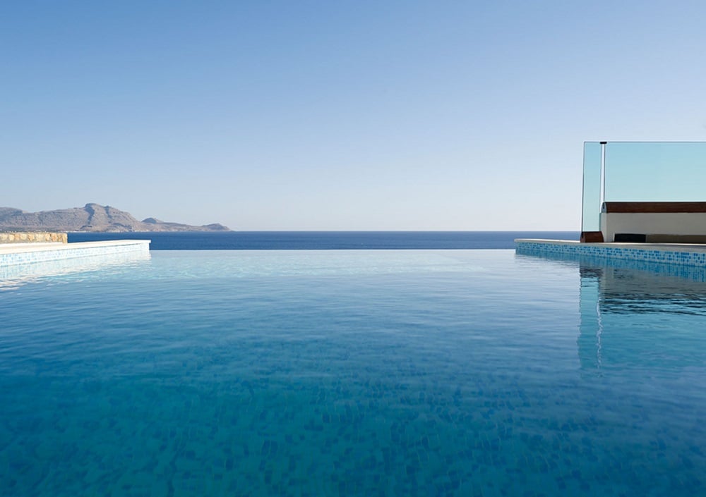 An infinity pool stretches out towards the horizon, seamlessly merging with the calm sea. The clear blue water reflects the sky, creating a tranquil setting with unobstructed views of the distant mountains and coastline.
