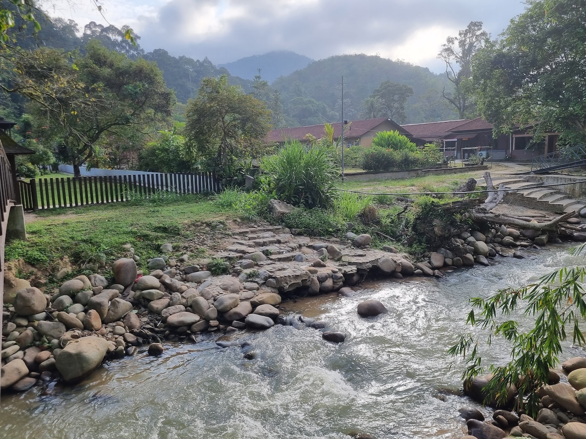 A river gently flows past smooth stones, framed by lush greenery and trees. A simple landscape features a wooden structure in the background, with a sloped roof visible. The serenity of the environment is enhanced by the nearby mountains, contributing to a peaceful retreat setting.