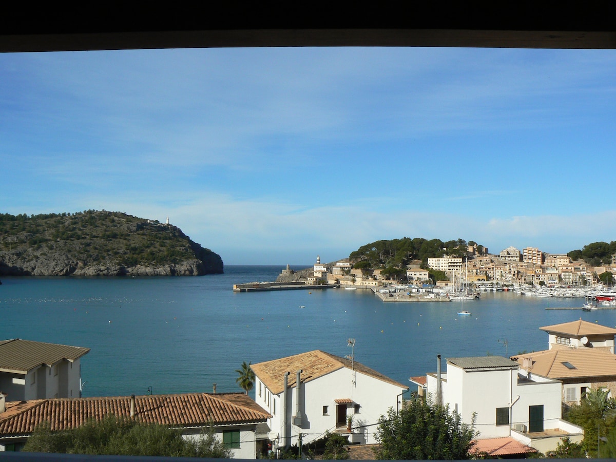 A scenic view of the coastline is captured from a window, revealing a tranquil harbor with boats anchored. Nearby, a lighthouse and charming village are visible against a backdrop of rolling hills under a clear blue sky.