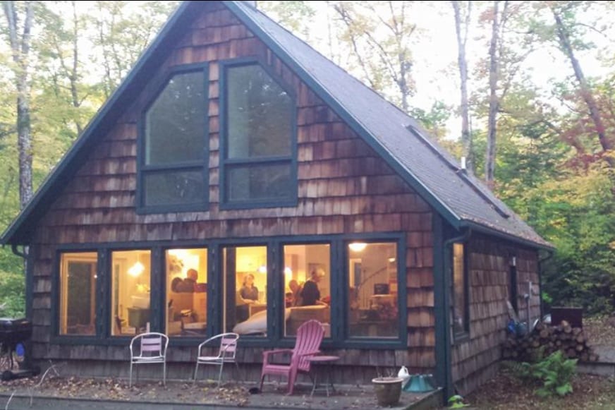 The chalet-style cabin features a distinctive wooden exterior adorned with numerous windows, allowing warm light to spill out. The surrounding trees create a natural backdrop, enhancing the serene atmosphere of the retreat. Two outdoor chairs are visible in the foreground.