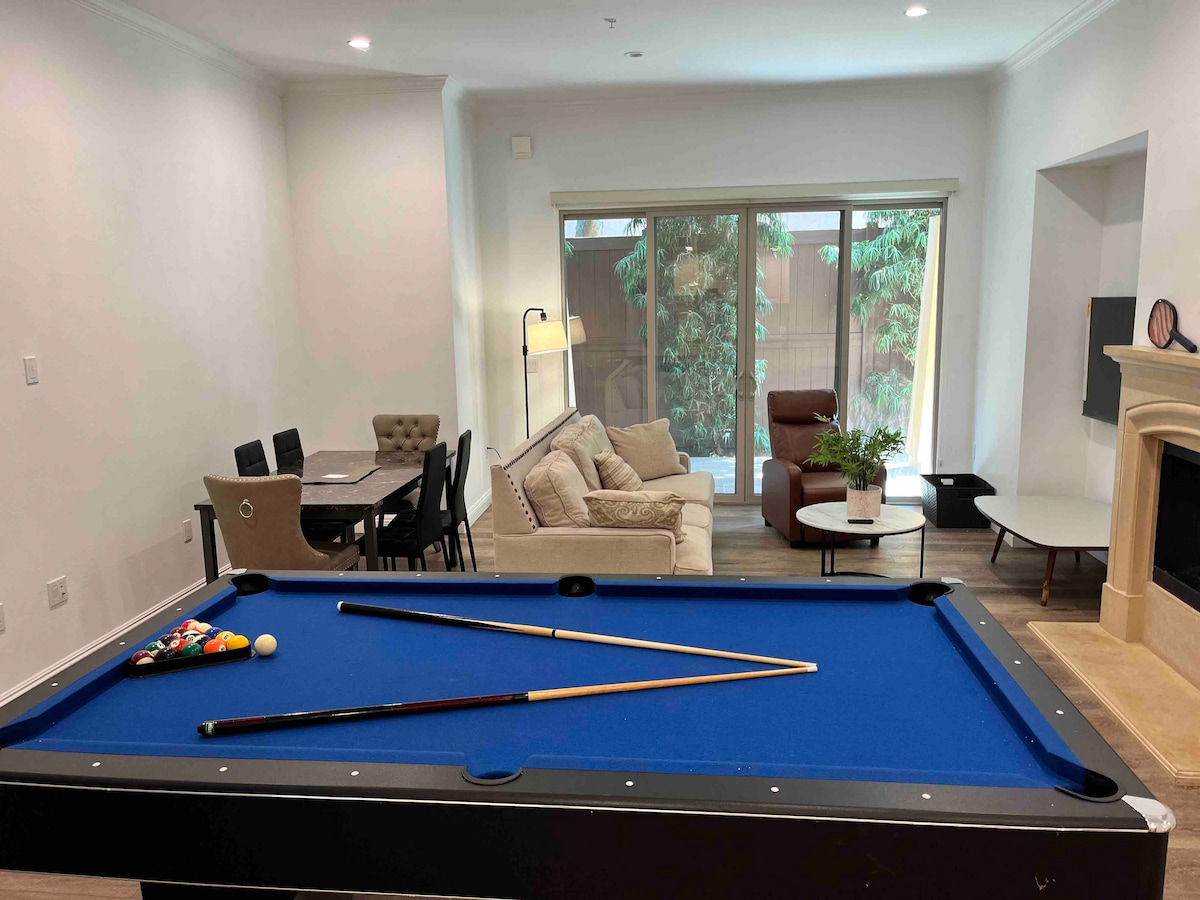 A spacious game area is highlighted by a standard pool table with a blue felt surface and two cues resting on it. In the background, a cozy seating arrangement includes a light-colored sofa and a dining table with four chairs, accompanied by large windows offering views of greenery.