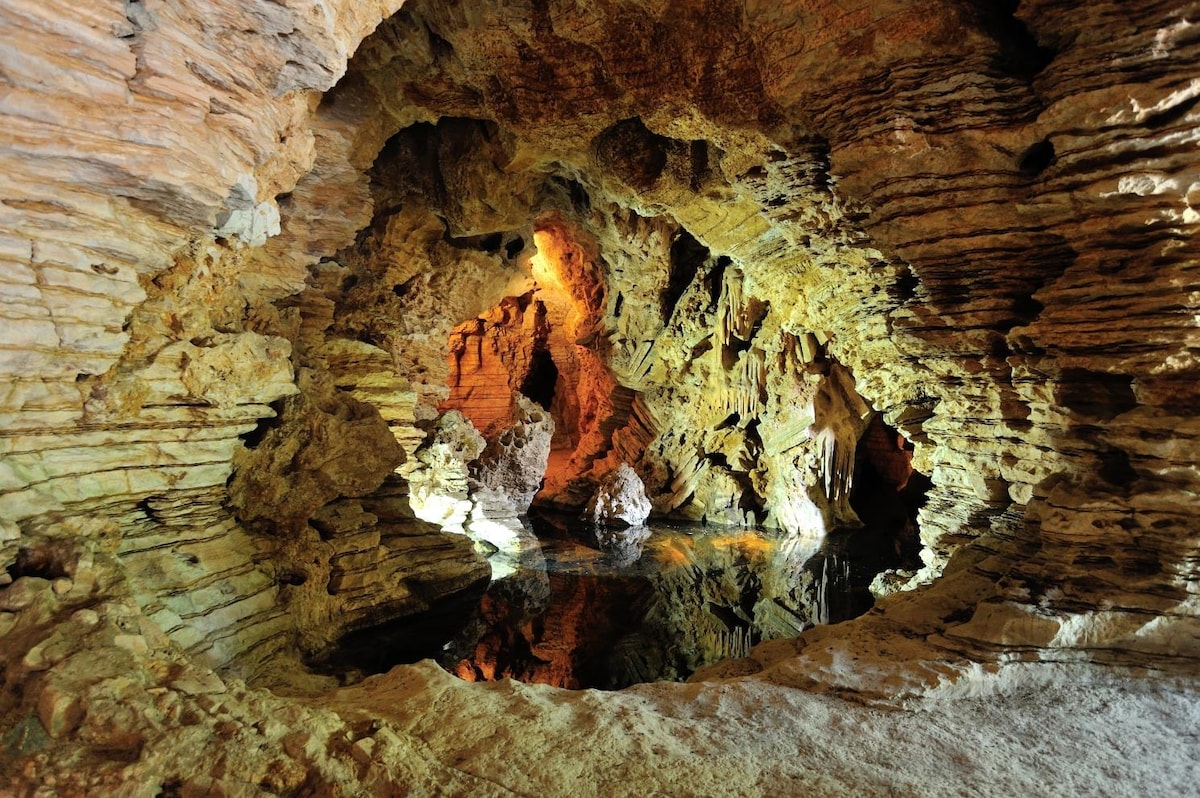A network of artificial caves features rugged rock formations and a tranquil water surface reflecting warm light. Textured stone walls create an intricate environment, highlighted by natural color variations. Pockets of illumination enhance the cave’s depth and unique characteristics, making it an intriguing spot for exploration.