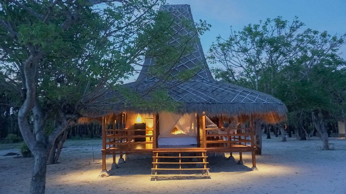 A traditional Sumba-inspired villa features a thatched roof and wooden structure elevated on stilts. Soft lighting highlights the open space beneath, where a spacious bed is visible. Surrounding trees provide shade, creating a serene environment, while sandy grounds are seen in the foreground.