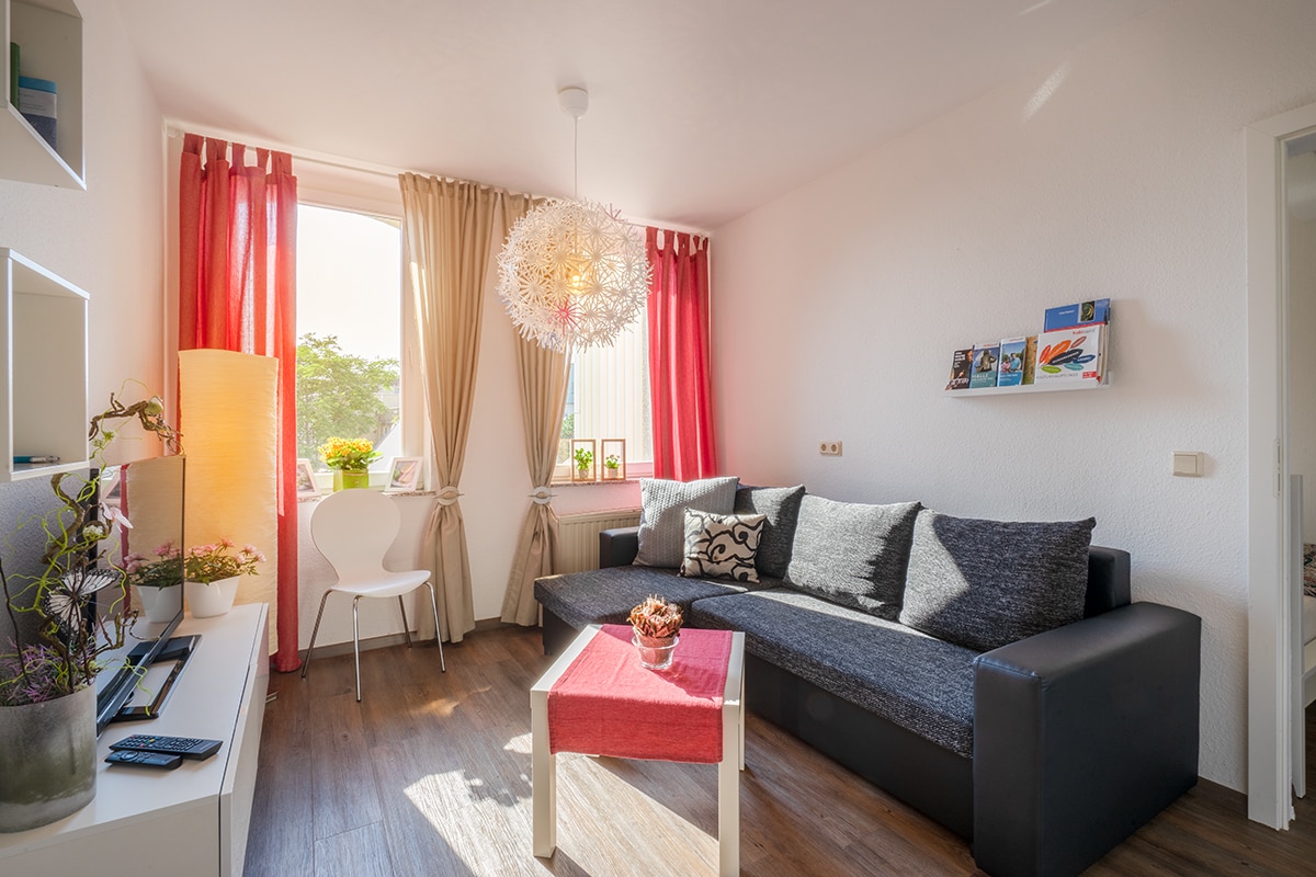 A bright living area is shown, featuring large windows covered by sheer red and white curtains. A dark sectional sofa rests beside a small red accent table. Natural light illuminates decorative elements, including a floor lamp and wall-mounted shelves with books.