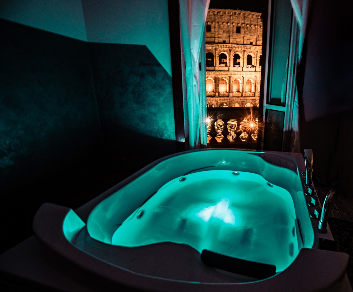 A spacious jacuzzi is positioned near a window, displaying a view of the illuminated Colosseum at night. Soft blue lighting enhances the jacuzzi environment, creating a serene atmosphere that complements the external view.