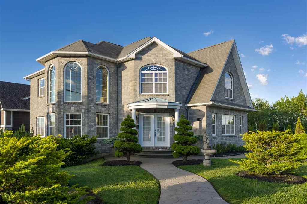 A stately two-story home characterized by its stone façade and multiple large arched windows. The entrance is framed by manicured shrubs and a stone pathway, leading to a welcoming front door. Lush green grass surrounds the house, complemented by well-maintained landscaping.
