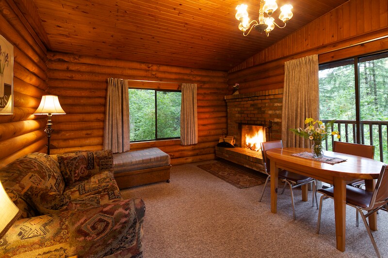 The living area features a rustic wood-paneled interior with a cozy seating arrangement, including a patterned sofa and a matching ottoman. A dining table with chairs sits adjacent to a stone fireplace, where a fire is lit. Large windows allow natural light to fill the space.