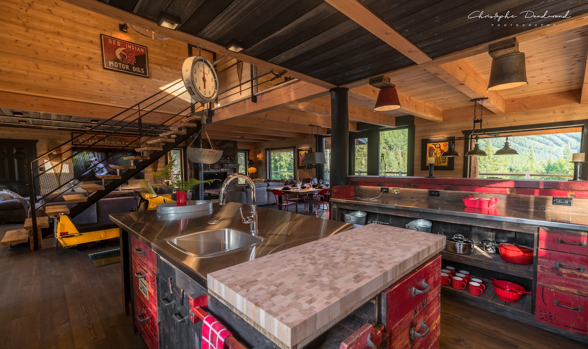An open kitchen features stainless steel countertops and rustic wooden cabinets with a vibrant red finish. Large windows provide natural light and scenic views of the surrounding nature. A staircase is visible in the background, leading to the upper level.