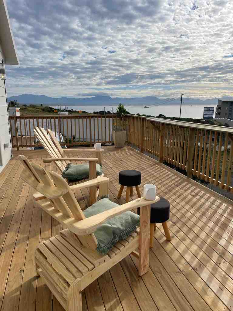 A wooden deck overlooks the ocean, featuring two spacious lounge chairs and two small tables. Soft cushions are placed on the chairs, adding comfort. The deck is bathed in natural light, with distant mountains visible under a partly cloudy sky.