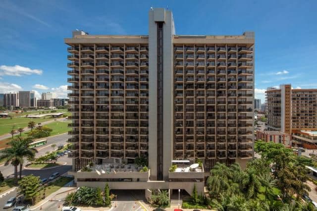 The hotel building is portrayed from an elevated angle, showcasing its modern, multi-story structure with numerous balconies. Surrounding palm trees and greenery frame the base, enhancing the urban setting. The sky is clear, allowing ample natural light to highlight the architecture.