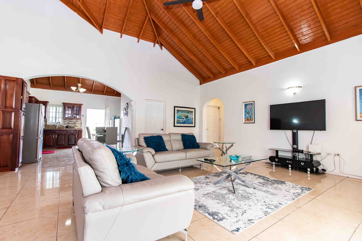 A spacious living area is characterized by a high, wooden-beamed ceiling and glossy tile flooring. Two light-colored sofas are arranged around a glass coffee table, with a vibrant area rug beneath. A large wall-mounted television and tasteful artwork are featured, enhancing the room's inviting atmosphere.