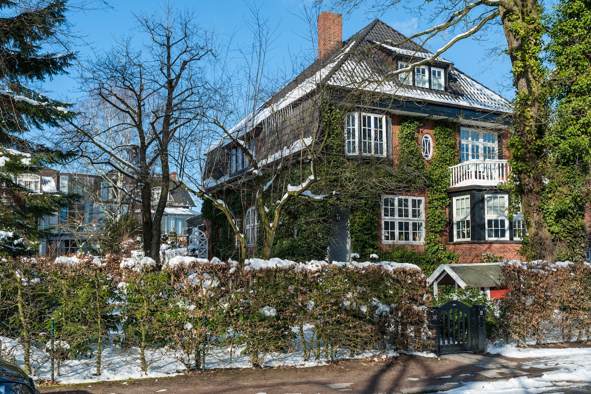 The historic Jugendstil house is framed by lush greenery and a freshly fallen snow. Its charming façade features multiple windows and a balcony on the upper level, reflecting the architectural style of the early 20th century. A tree-lined driveway leads to the entrance.