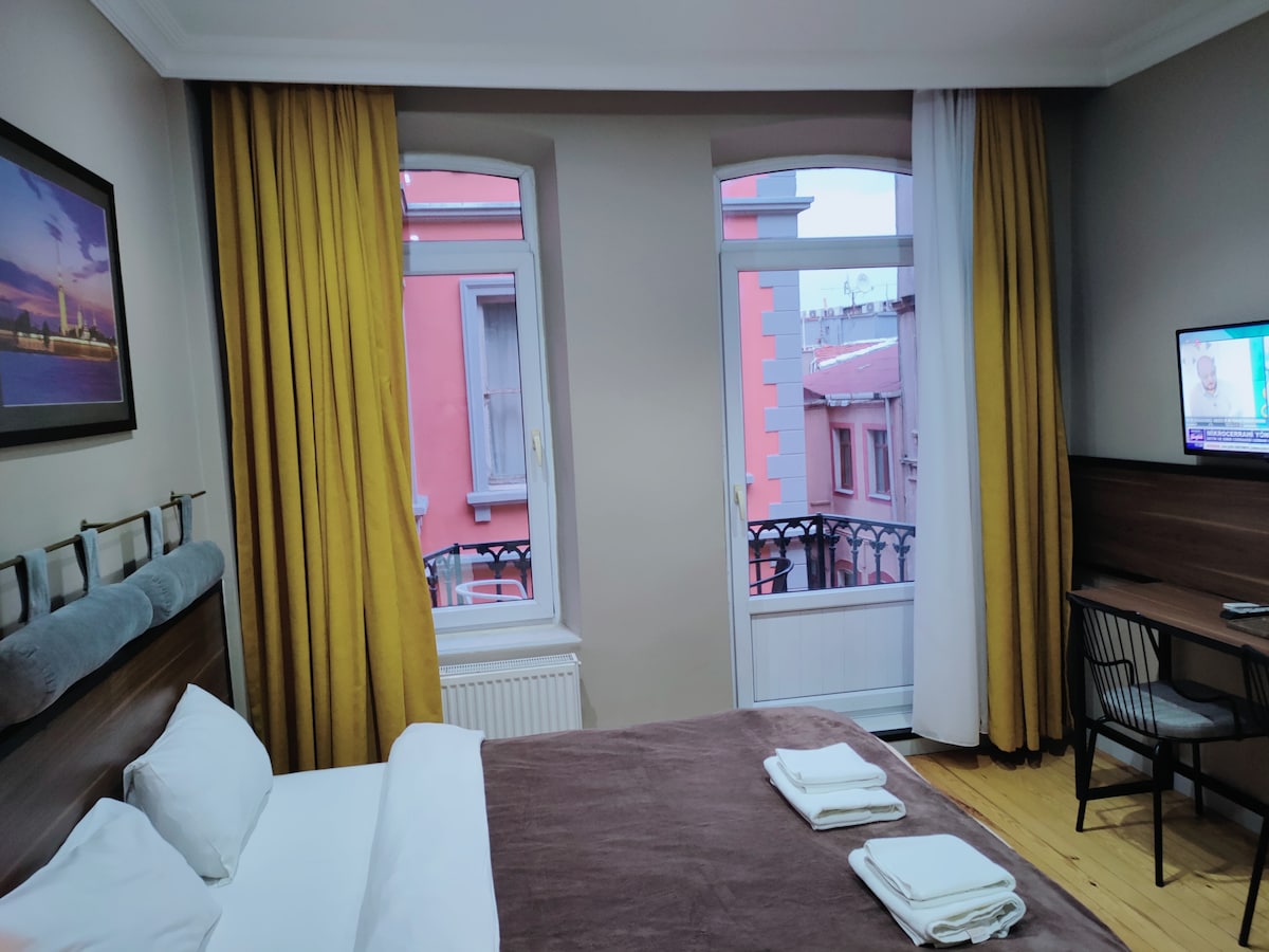 A well-lit room features a spacious bed with neatly arranged towels on a brown blanket. Large windows are dressed with golden curtains, offering views of the balcony. A small desk with a chair and a television are positioned against the wall.