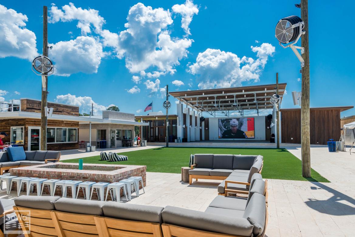 An outdoor amphitheater is presented, featuring a large screen and a stage, with a spacious seating area. Comfortable seating arrangements in a modern design are arranged on a lush green lawn, surrounded by a vibrant blue sky dotted with fluffy white clouds.