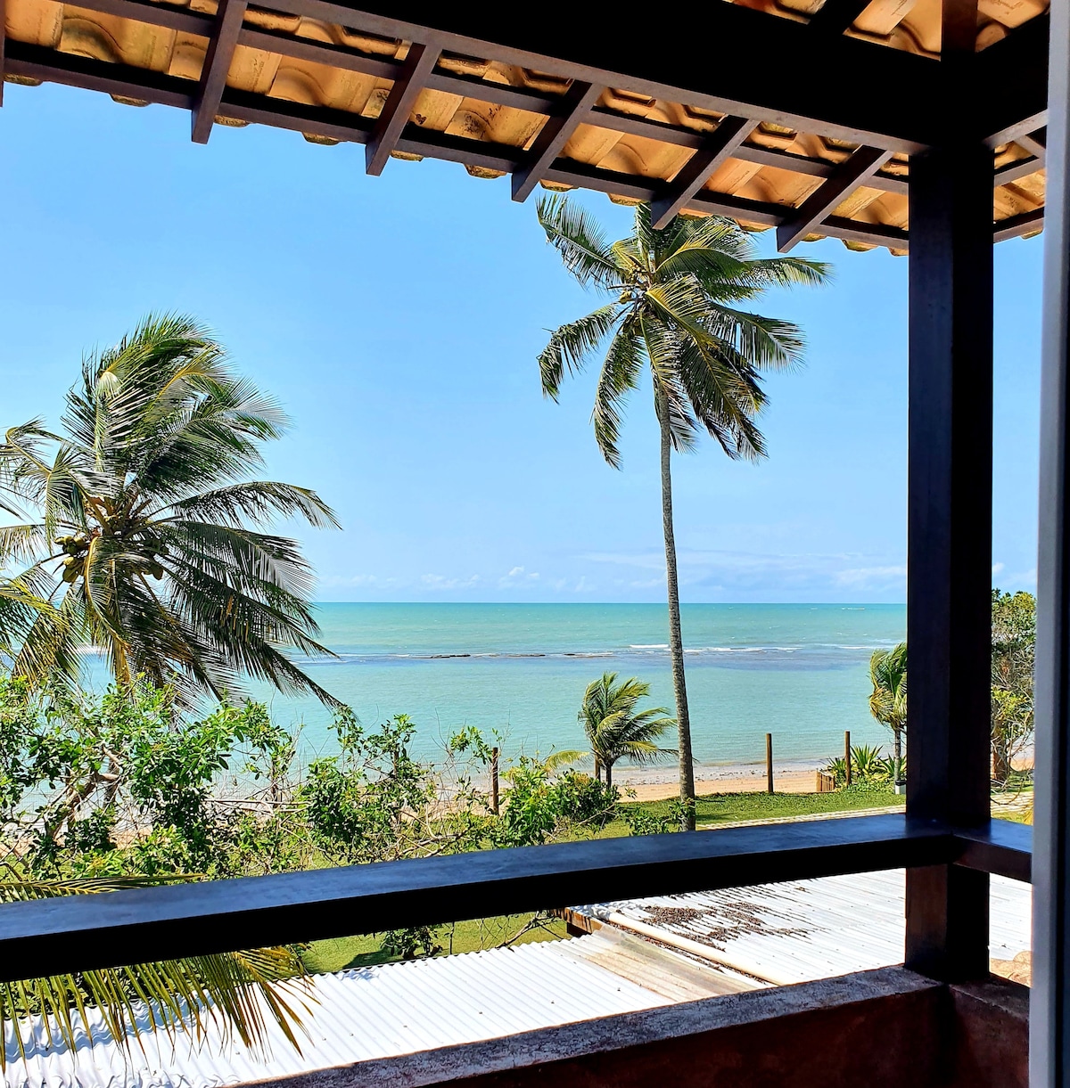 A view of the ocean is framed by wooden beams and a thatched roof, highlighting swaying palm trees against a clear blue sky. The calm water reflects natural light, creating a serene coastal atmosphere.