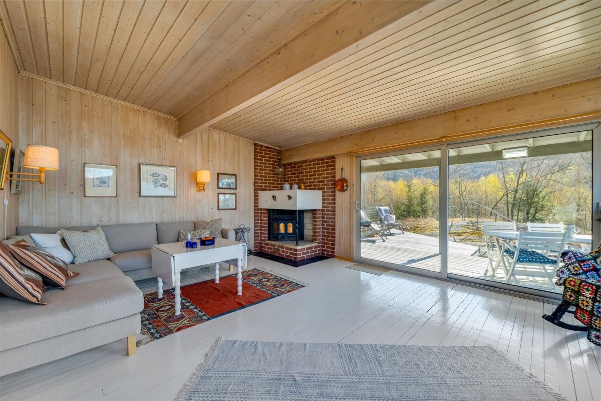 A warm living area features light wood paneling and a cozy fireplace, complemented by a soft grey sofa and rectangular coffee table. Large glass doors lead to a scenic outdoor terrace, while a vibrant rug and colorful quilt add comfort to the space.