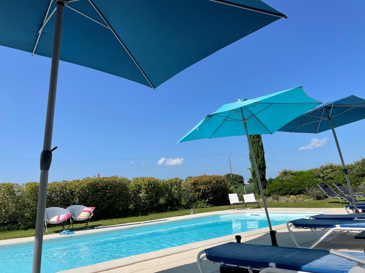 A serene swimming pool is visible beneath a clear blue sky, surrounded by lush greenery. Parasol shades provide comfort for guests relaxing on lounge chairs nearby, creating a peaceful outdoor setting.