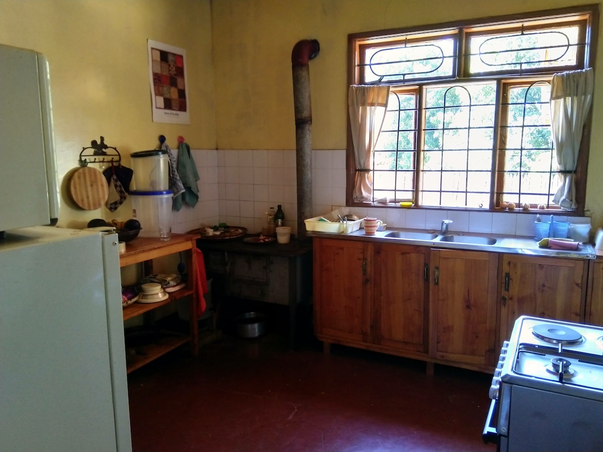 A functional kitchen equipped with wooden cabinetry and a stainless steel sink. Natural light enters from the large window adorned with light curtains. An arrangement of cooking utensils is visible on the countertop, along with a traditional stove and an adjacent wooden shelf storing various items.