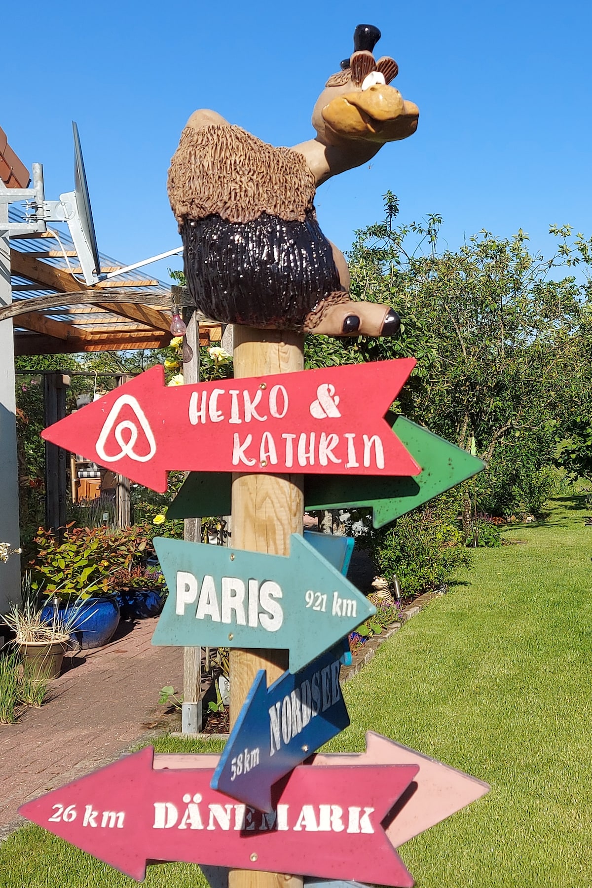 A colorful wooden signpost featuring directional arrows indicates various distances to locations such as Paris, Denmark, and Nordsee. A whimsical character is perched atop, adding a playful element to the outdoor garden area, surrounded by greenery and blooming flowers.