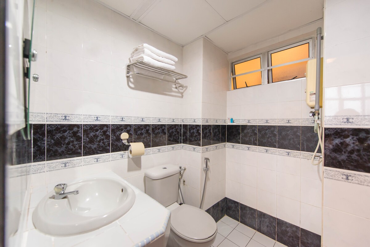 The bathroom features a compact layout with white and black tiled walls. A sink is positioned beside a toilet, and a shower area is visible. Towels are neatly arranged on a metal shelf, and natural light enters through a window with an orange hue.