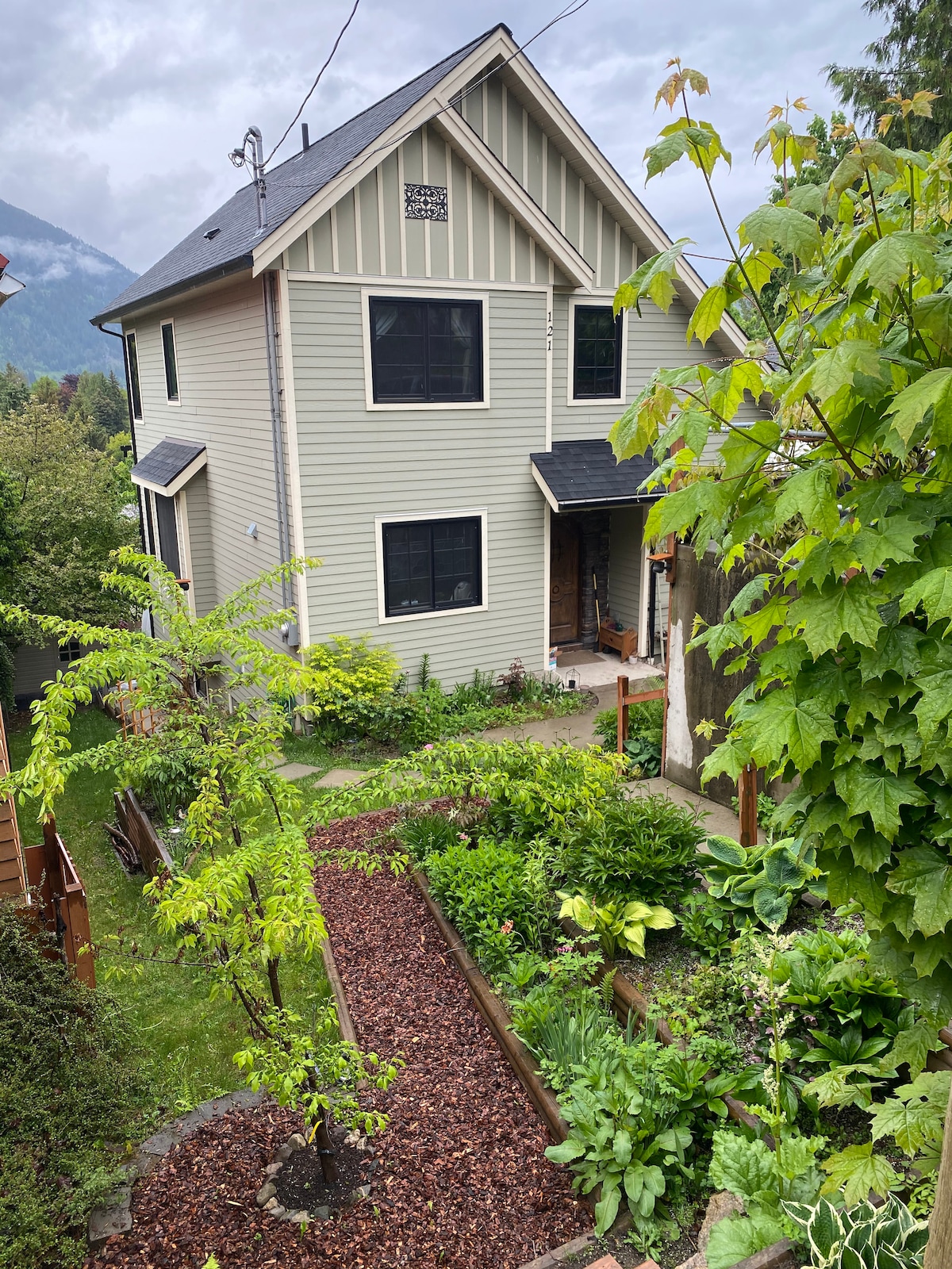 The exterior of a three-story house is visible, featuring a light-colored siding and dark windows. A landscaped garden with mulch pathways and various plants surrounds the home. The entrance is framed by greenery, providing a natural and inviting atmosphere.