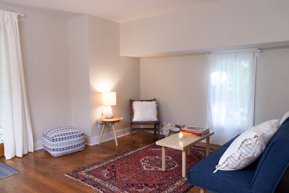 The cozy studio features a textured area rug placed on hardwood floors. A comfortable blue sofa and a small table invite relaxation. Soft white curtains frame a window, while a lamp offers gentle illumination beside a wooden chair adorned with a plush cushion.