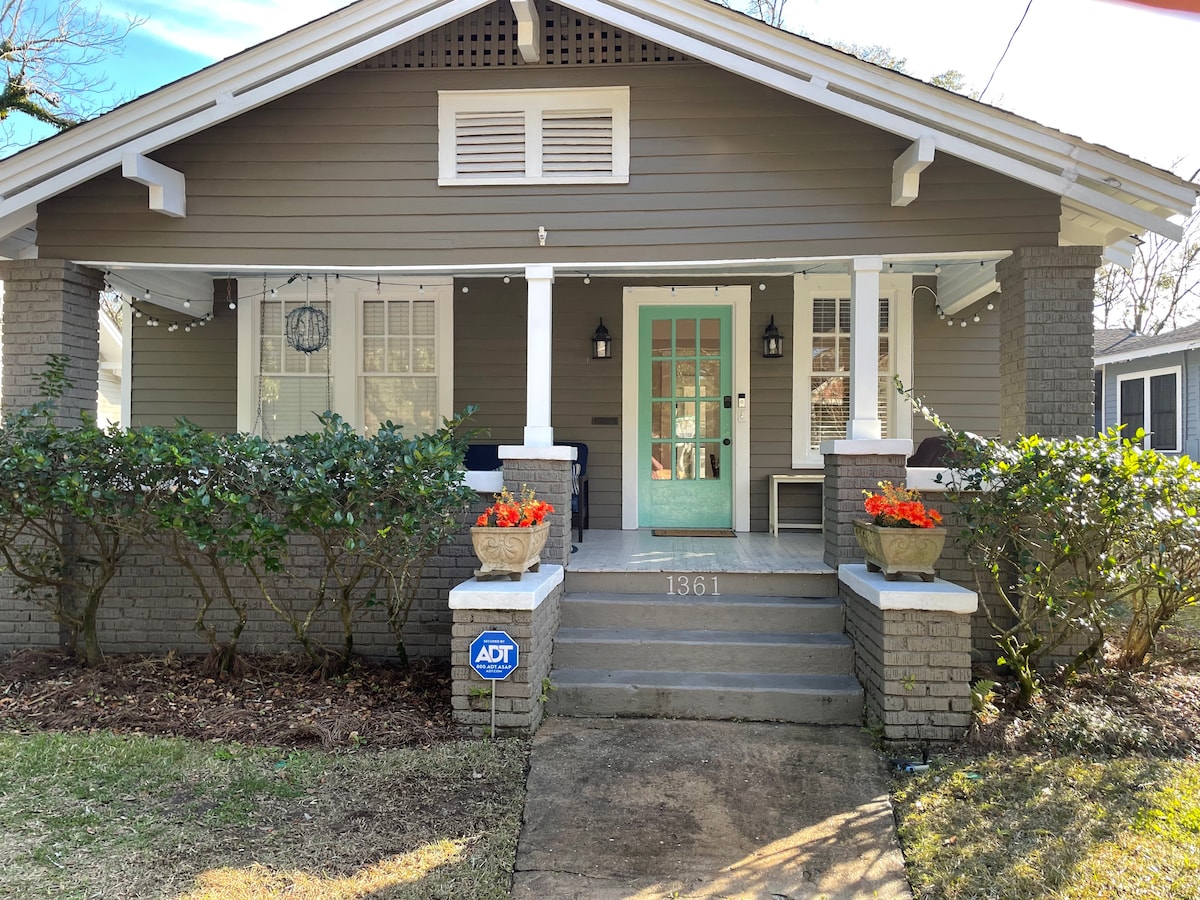The charming bungalow features a welcoming front porch with light green doors and a symmetrical layout. Potted flowers add a splash of color to the entrance. The porch is framed by neatly trimmed bushes and covered by a sloping roof with decorative details.