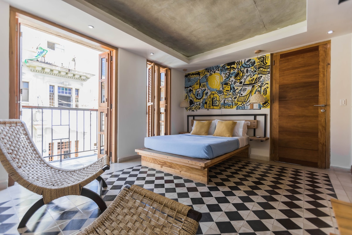 The room features a spacious bed with neutral linens, set against a vibrant hand-painted wall. Natural light floods in through two open wooden windows, overlooking a lively street. A woven chair adds a touch of comfort to the modern tile floor.