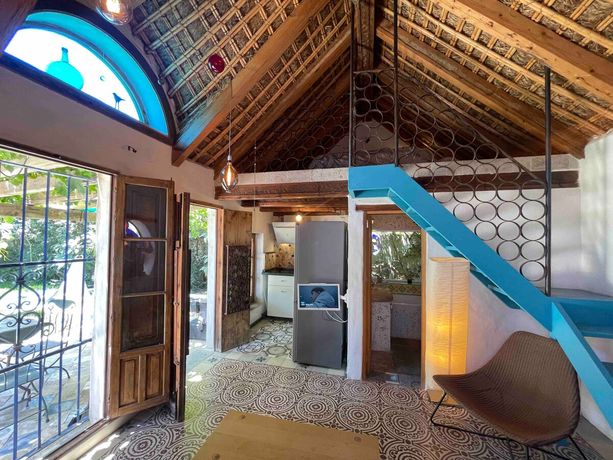 An interior space featuring high natural straw ceilings and decorative tile flooring. Open wooden doors provide access to the terrace, while a spiral staircase leads to a loft area. A kitchenette with appliances is visible in the background, enhancing the functional layout.