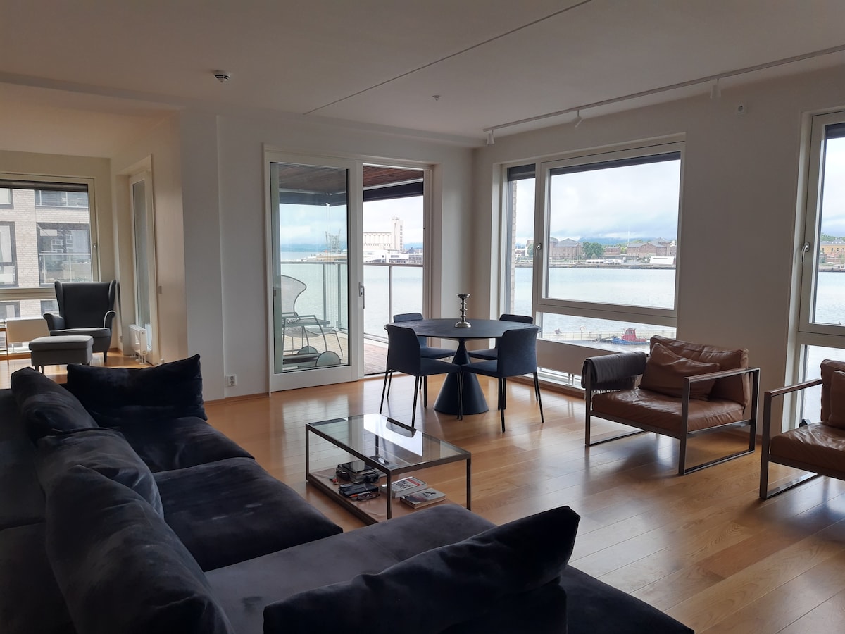 The spacious living area features large windows that invite natural light and provide a view of the waterfront. A comfortable sofa, a glass coffee table, and two armchairs are arranged for relaxation. A dining table for four is positioned by the windows.