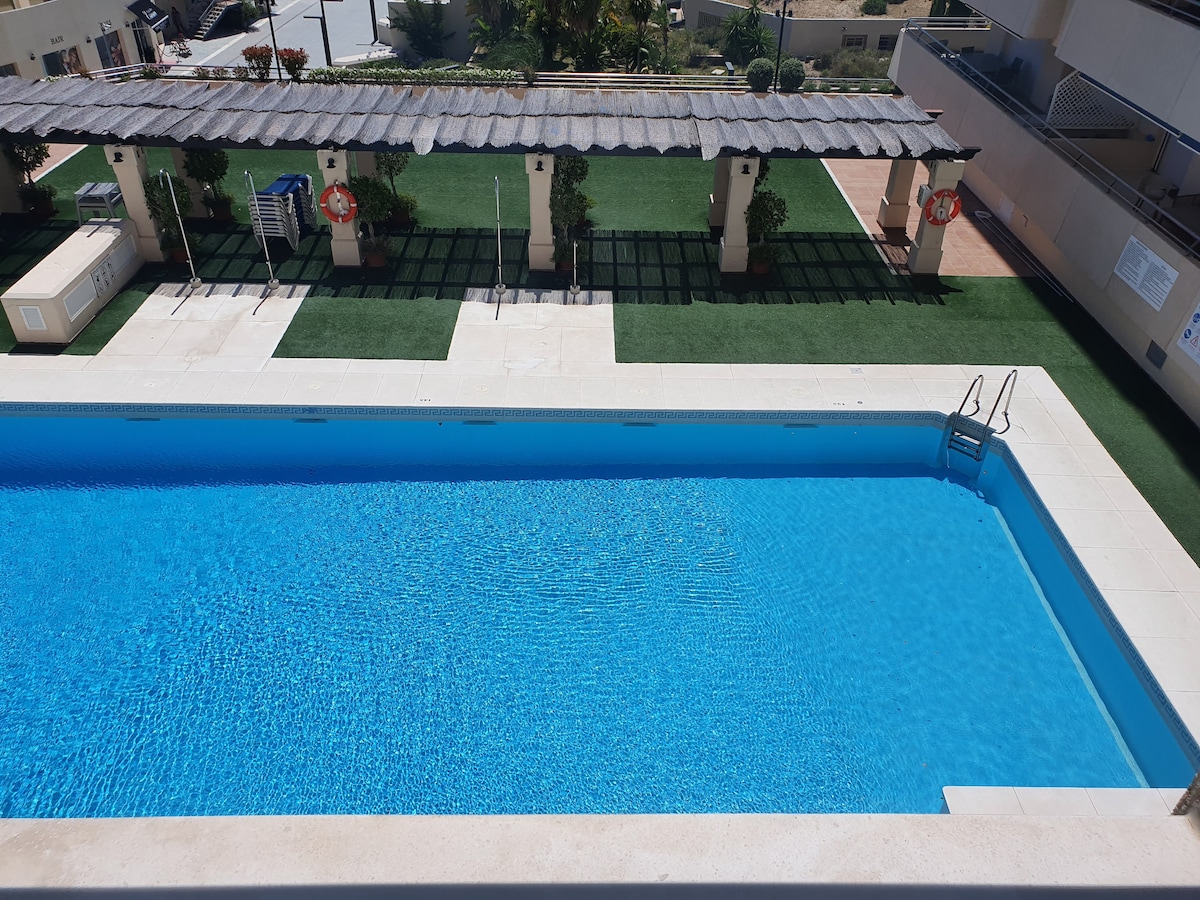 A rectangular swimming pool is surrounded by well-maintained greenery and shaded areas. Blue water glistens under sunlight, while steps at one end provide access to the pool. Lounge chairs and structured canopies are visible in the background, contributing to a relaxed outdoor environment.