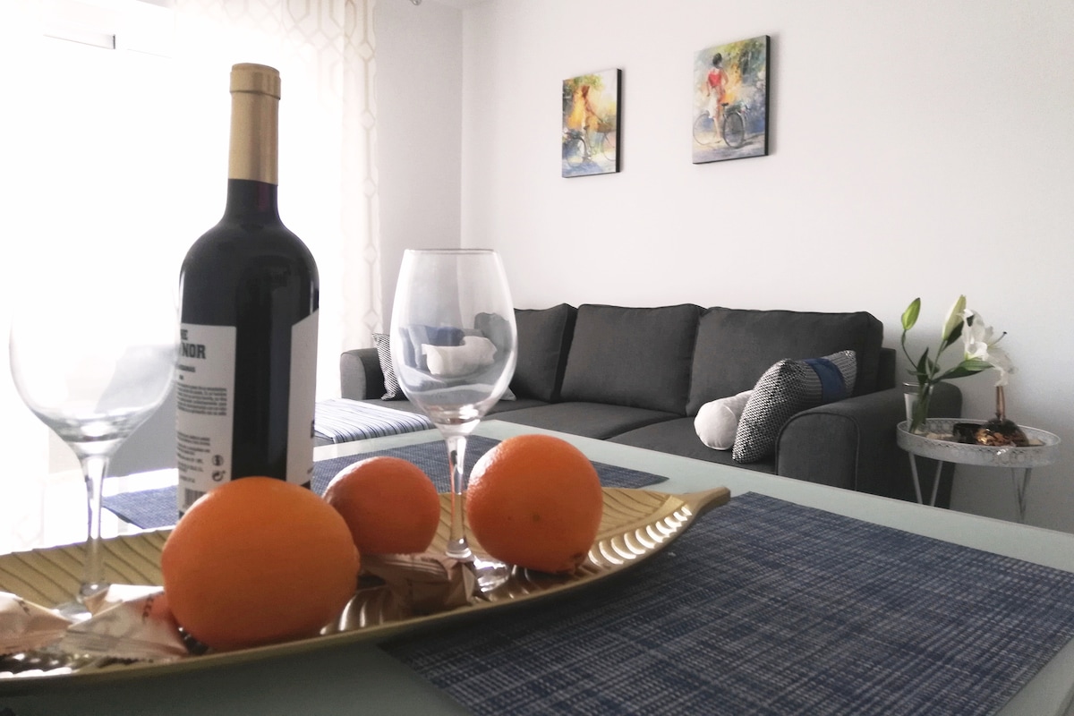 A dining table is set with a gold tray holding oranges and chocolates, accompanied by two wine glasses and a bottle of wine. In the background, a gray sofa is placed against a wall adorned with two small framed artworks, while natural light illuminates the space.