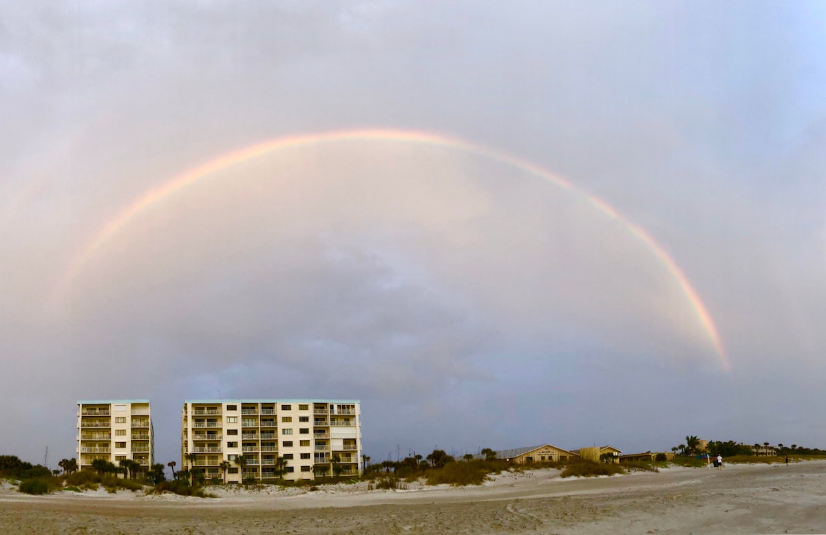A gentle rainbow arches over the beach, illuminating the sky. Two multi-story buildings stand in the foreground, framed by low coastal vegetation and sandy terrain. The scene captures a peaceful coastal atmosphere, suggesting proximity to the beach.