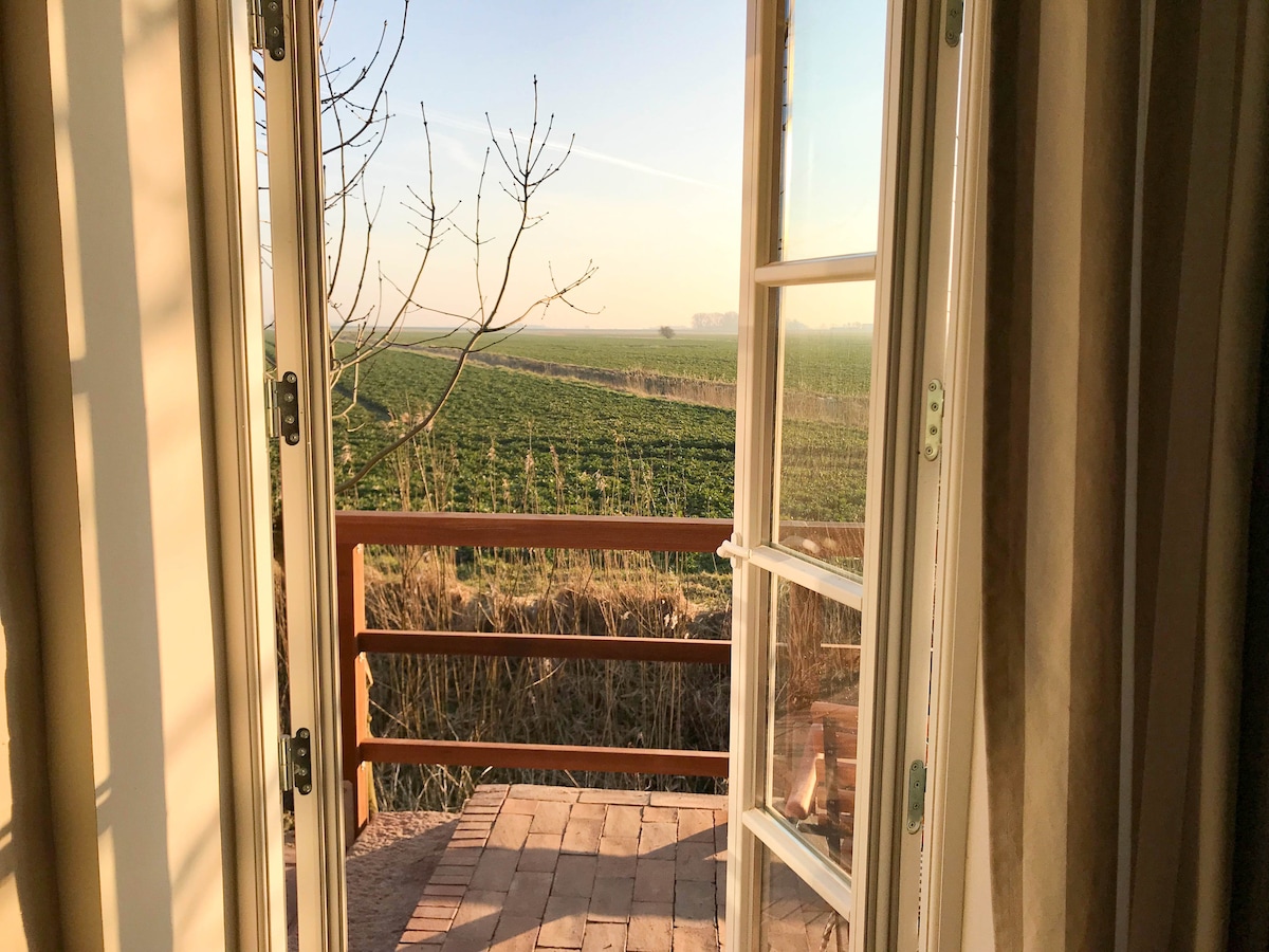 Double doors open onto a wooden balcony, revealing expansive views of green fields bathed in soft morning light. A tree branch is visible in the foreground, while the horizon stretches with gentle farmland.