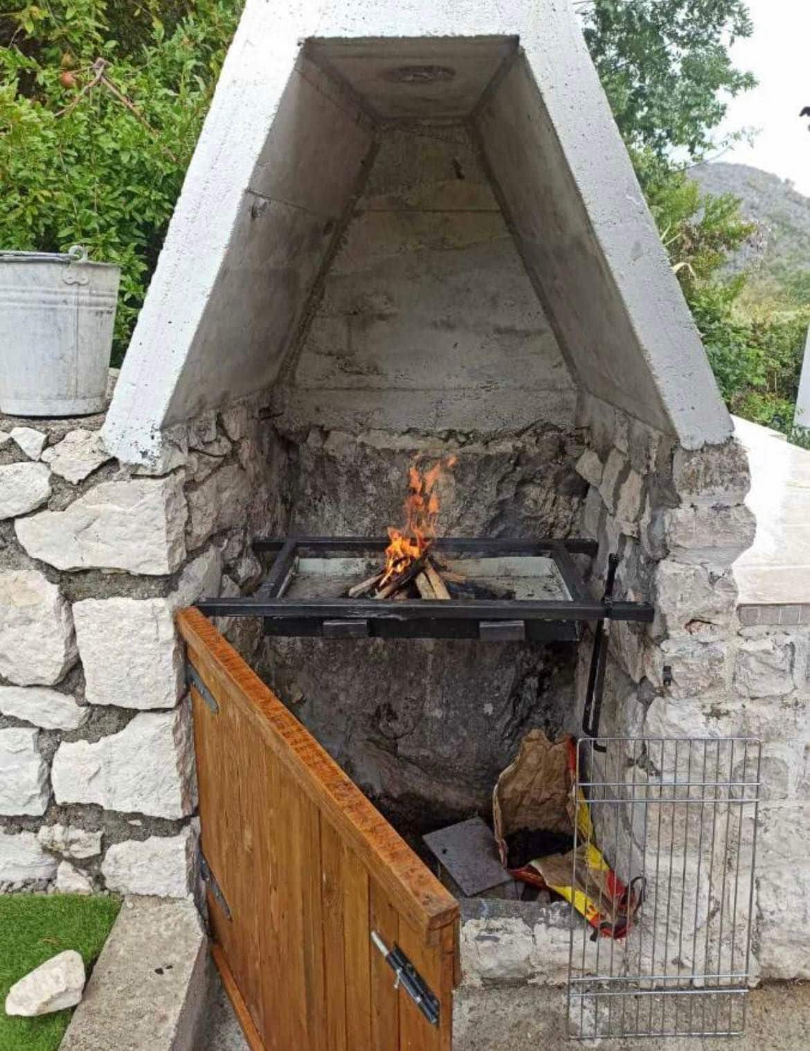 A stone-built outdoor fireplace is surrounded by natural greenery. A fire is being contained within a metal frame, providing a warm glow. The fireplace design includes a wooden door and a grilling rack, offering a functional space for outdoor cooking.